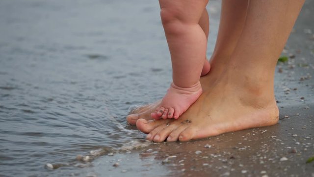 Little baby child feet stand on mother feet at seashore, first feeling of sea waves