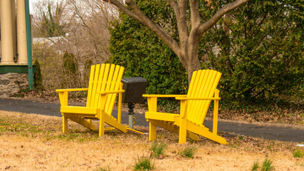 Two Bright Yellow Wooden Lawn Chairs on a Dead Lawn With a Driveway in the Background