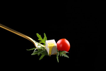 Feta cube cheese with arugula and ripe cherry tomatoes salad on a golden fork, with a flowing drop of olive oil, on a black background.