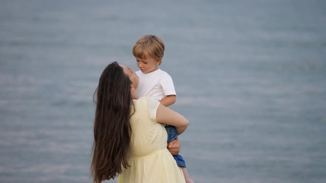 Mother standing at sea shore hold little child, lift boy and kiss son, sea waves