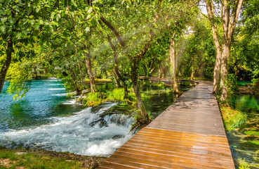 Wooden footpath over river in forest of Krka National Park, Croatia. Beautiful scene with trees, water and sunrays. - obrazy, fototapety, plakaty