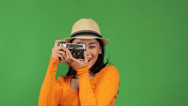 Pretty asian woman in stylish outfit actively taking photos through a retro camera on green background. Hobbies, leisure, favorite pastime. Fun, relaxation, photoset. Female portrait