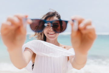 Joyful tanned girl playfully posing with sunglasses on sea background. Outdoor photo of pretty young woman fooling around at beach.
