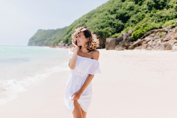 Fototapeta premium Graceful woman in dress looking at sea with cheerful smile. Outdoor photo of gorgeous blonde lady spending time at tropical island.