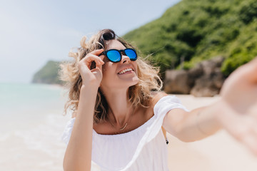 Cheerful girl with tanned skin making selfie at tropical island. Outdoor photo of ecstatic young woman in trendy sunglasses taking picture of herself at sandy beach.