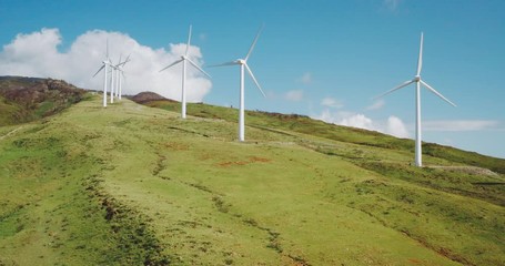 Aerial view of a row of wind turbines spinning on a hill with green grass and blue skies, new types of energy generation for a cleaner and greener renewable energy future