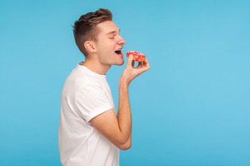 Junk food. Side view of delighted man in white t-shirt eating donut with expression of big...