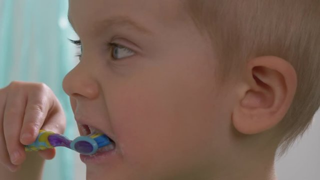 Child (toddler, little boy) is brushing his teeth . Taking care of teeth, cleaning teeth using fluoride toothpaste in a bathroom, dental care concept. Closeup kid's face, looking at camera