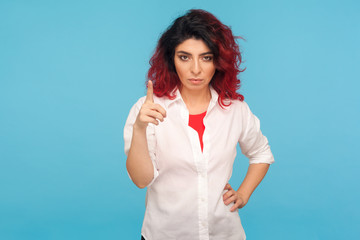 Be careful! Portrait of bossy displeased hipster woman with fancy red hair in white shirt holding finger up in admonishing gesture, warning about danger. indoor studio shot isolated on blue background