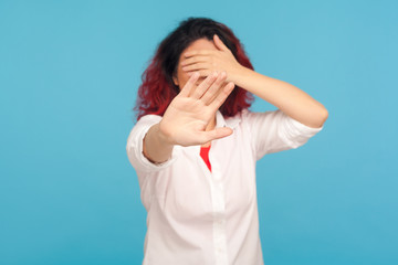 Portrait of embarrassed woman showing stop gesture, covering eyes with hands so not to watch forbidden shameful content, pretending to not see problems. indoor studio shot isolated on blue background