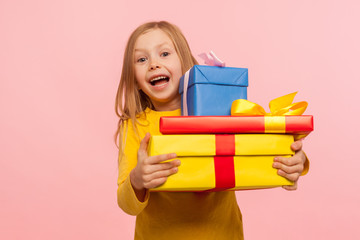 Delighted amazed little girl embracing lot of gift boxes, looking at camera with surprise and sincere childish happiness, shocked by many birthday presents. indoor studio shot, pink background