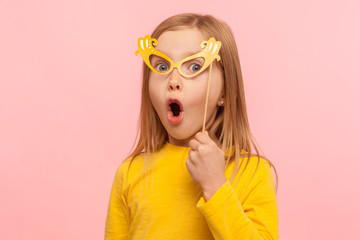 Surprised little girl covering eyes with paper glasses and sticking out tongue, naughty child...