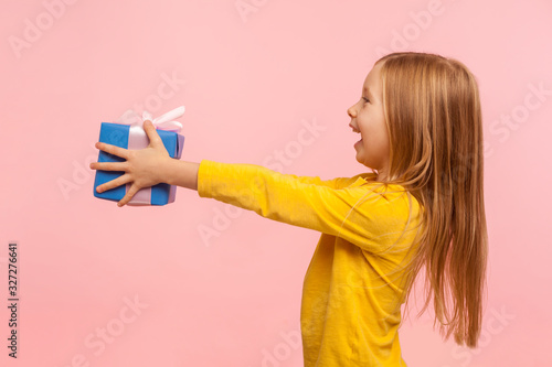 Take it! Side view of generous kind adorable little girl giving present and congratulating on birthday, mother's day, offering gift box for charity. indoor studio shot isolated on pink background