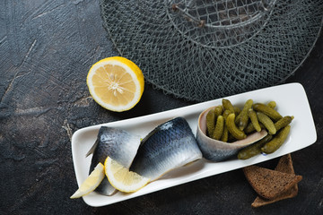White plate with herring fillets and pickles, elevated view over dark brown stone surface with a metal fish cage, studio shot