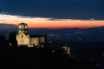 View of St. Francis papal church in Assisi at night, with city lights on the background