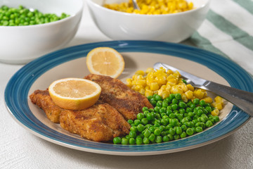 Golden batter deep fried fish fillet, served with green peas, sweet corn, and slices of lemon close up on a plate on white background