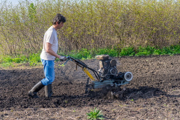 Man in wellingtons with cultivator ploughing ground in sunny day. Farmer plowing kitchen-garden in suburb. Land cultivation, soil tillage. Spring work in garden. Gardening concept