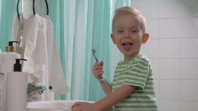 Child (toddler, little boy) is brushing his teeth . Taking care of teeth, cleaning teeth using fluoride toothpaste in a bathroom, dental care concept. Closeup kid's face, looking at camera