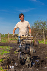 Man in wellingtons with cultivator ploughing ground in sunny day. Farmer plowing kitchen-garden in suburb. Land cultivation, soil tillage. Spring work in garden. Gardening concept