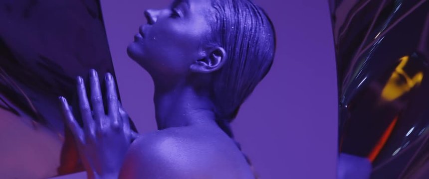 sexy young model with braid tail posing in neon UV light looking at camera, purple and blue portrait of 20s woman or girl, creative makeup, face skin and hair covered with glitter metallic shine paint