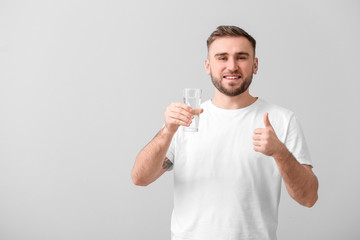 Young man with glass of water showing thumb-up on light background