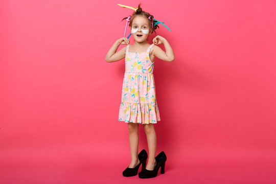Indoor shot of little girl in big mother's shoes, charming female kid isolated over pink background, excited little child with curlers and patches under eyes, full length portrait of charming kid.