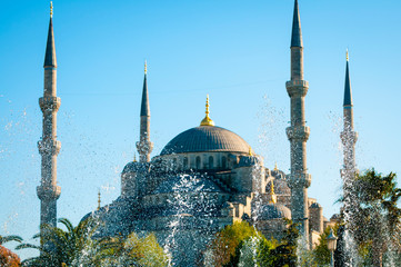Sunny view of the dome and minarets of the Blue Mosque through fountain spray under blue sky in...