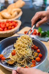 closeup of a chef forking up spaghetti Vongole with roma tomatoes and mussels in a pan - with a knife and cutboard with tomatoes and bread in the background