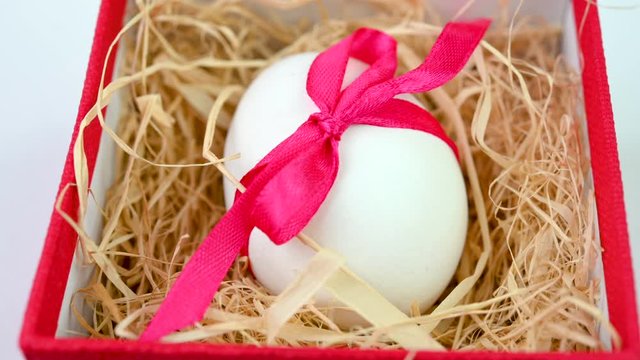 white Easter egg decorated with a red ribbon lies in a small gift box on a straw. Easter decorations. Easter concept background.
