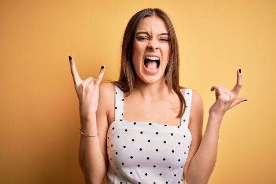 Young beautiful brunette woman wearing casual dress standing over yellow background shouting with crazy expression doing rock symbol with hands up. Music star. Heavy concept.