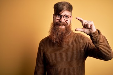 Handsome Irish redhead man with beard wearing glasses and winter sweater over yellow background smiling and confident gesturing with hand doing small size sign with fingers looking and the camera