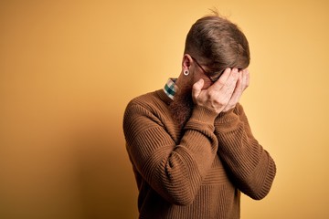 Handsome Irish redhead man with beard wearing glasses and winter sweater over yellow background with sad expression covering face with hands while crying. Depression concept.