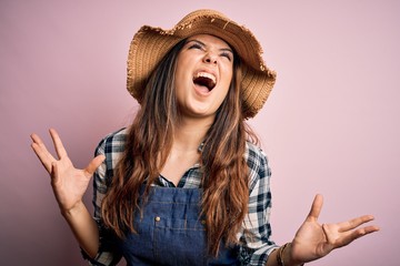 Young beautiful brunette farmer woman wearing apron and hat over pink background crazy and mad shouting and yelling with aggressive expression and arms raised. Frustration concept.