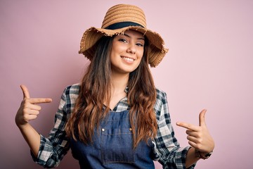 Young beautiful brunette farmer woman wearing apron and hat over pink background looking confident...