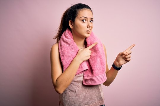 Young beautiful brunette sportswoman wearing sportswear and towel over pink background Pointing aside worried and nervous with both hands, concerned and surprised expression