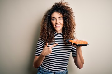 Young beautiful woman with curly hair and piercing holding tray with fresh sushi very happy pointing with hand and finger