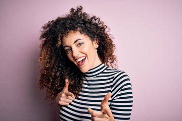 Young beautiful woman with curly hair and piercing wearing casual striped t-shirt pointing fingers...