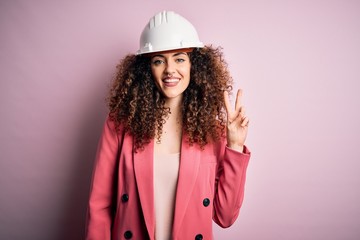 Young beautiful architect woman with curly hair wearing safety helmet over pink background smiling with happy face winking at the camera doing victory sign. Number two.