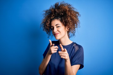 Young beautiful woman with curly hair and piercing wearing casual blue t-shirt pointing fingers to camera with happy and funny face. Good energy and vibes.