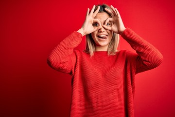 Young beautiful blonde woman wearing casual sweater over red isolated background doing ok gesture like binoculars sticking tongue out, eyes looking through fingers. Crazy expression.