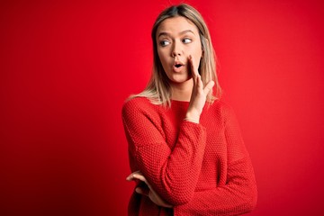 Young beautiful blonde woman wearing casual sweater over red isolated background hand on mouth telling secret rumor, whispering malicious talk conversation