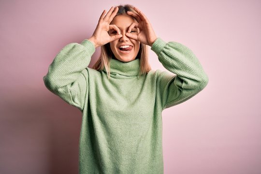 Young beautiful blonde woman wearing winter wool sweater over pink isolated background doing ok gesture like binoculars sticking tongue out, eyes looking through fingers. Crazy expression.