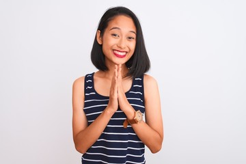 Young chinese woman wearing striped t-shirt standing over isolated white background praying with hands together asking for forgiveness smiling confident.
