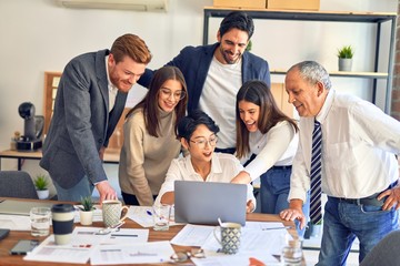 Group of business workers smiling happy and confident. One of them sitting and partners standing around. Working together with smile on face looking at the laptop at the office