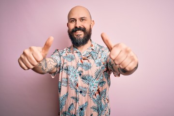 Fototapeta premium Handsome bald man with beard and tattoo wearing casual floral shirt over pink background approving doing positive gesture with hand, thumbs up smiling and happy for success. Winner gesture.