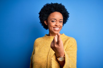 Young beautiful African American afro woman with curly hair wearing yellow casual sweater Beckoning come here gesture with hand inviting welcoming happy and smiling