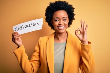 African American afro businesswoman with curly hair holding paper with capitalism message doing ok sign with fingers, excellent symbol - obrazy, fototapety, plakaty