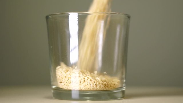 Raw sesame seeds in transparent glass with white reflection. Cereals pouring into an empty glass on a pastel background, close-up.