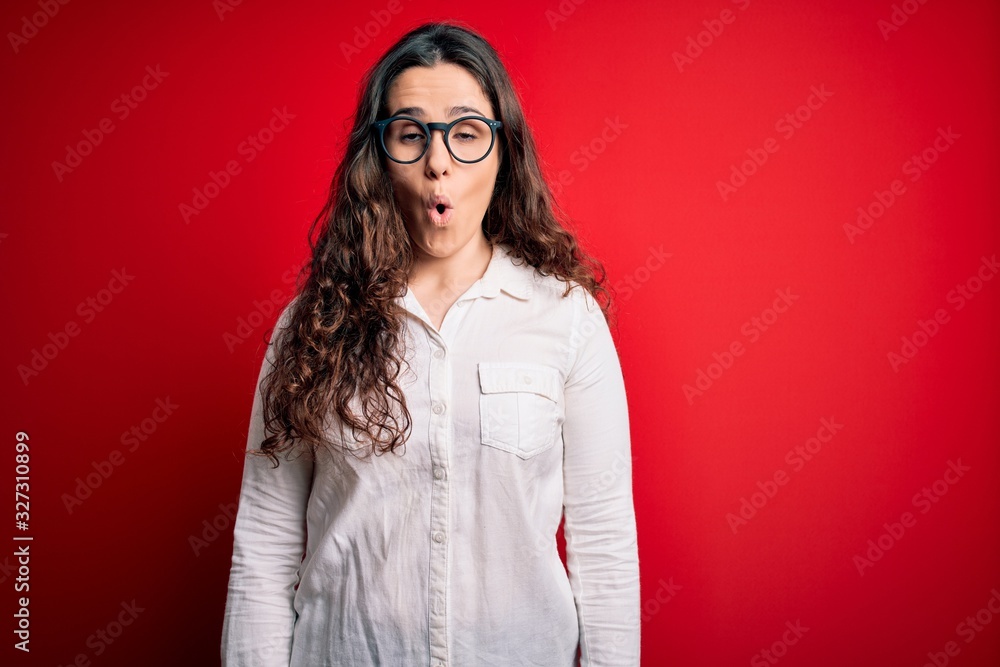 Sticker young beautiful woman with curly hair wearing shirt and glasses over red background afraid and shock