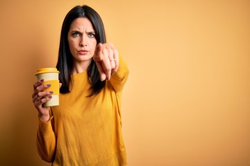 Young woman with blue eyes holding cup of coffee standing over yellow background pointing with finger to the camera and to you, hand sign, positive and confident gesture from the front